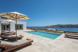 A poolside area with lounge chairs and an umbrella, overlooking a clear blue sea and a mountainous landscape. - Villa Sozon. Spacious villa with sea view 150 m from the beach. (Mykonos)