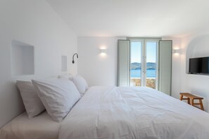 A bedroom with a bed, a small wooden stool, a TV, and a view of the sea through a door. - Villa Sozon. Spacious villa with sea view 150 m from the beach. (Mykonos)