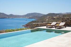 A swimming pool with lounge chairs, a stone wall, and a view of the sea and hills. - Villa Sozon. Spacious villa with sea view 150 m from the beach. (Mykonos)