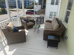 A covered porch with wicker furniture, a small table, and a view of the street and buildings. - Cute Bungalow in Cherry Grove, SC. Just a Chip Shot from ocean & beach access! (North Myrtle Beach)