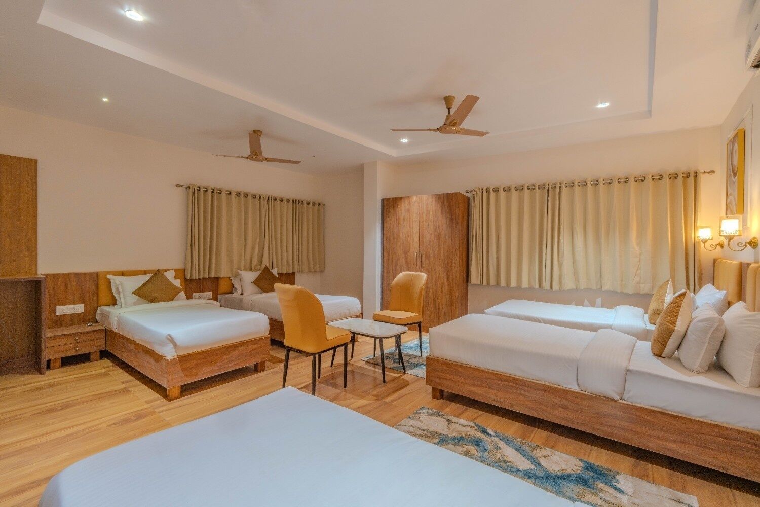 A hotel room with two single beds, a sofa bed, a small table, and a chair. There are ceiling fans and curtains on the windows.