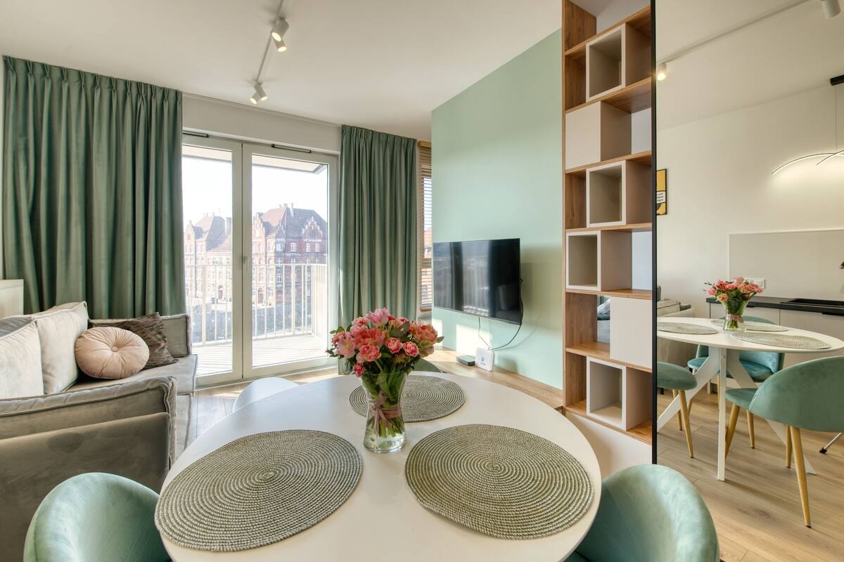 A modern living room with a sofa, a dining table with placemats, a TV, and a view of buildings through large windows.