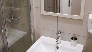 Egy fürdőszoba, melyben üvegfalas zuhanyzó, egy fehér mosdókagyló található, felette tükörrel, és alatta egy fa szekrény.