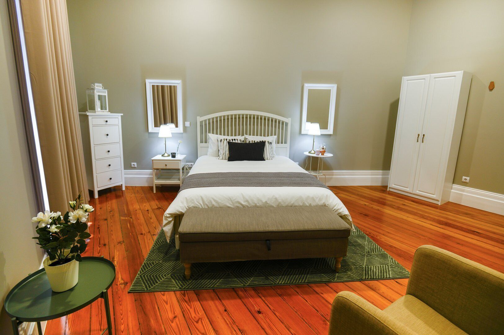 A bedroom with a large bed, two bedside tables with lamps, a dresser, a wardrobe, and a small table with a plant.