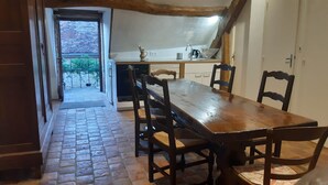 Een smalle gang die uitkomt op een eetgedeelte met een houten tafel en stoelen, een keuken op de achtergrond en een uitzicht door een glazen deur.
