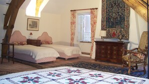 Een slaapkamer met een schuine kap, twee bedden met bijpassende hoofdeinden, een houten dressoir, een stoel en een groot wandtapijt.