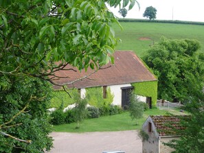 Uma casa de campo com telhado de telhas vermelhas, rodeada por vegetação exuberante e um campo verdejante.