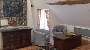 Een woonkamer met een donker houten kast, een lichtblauwe fauteuil, een kussen met dessin, een klein houten tafeltje en een groot bloementapijt aan de muur.