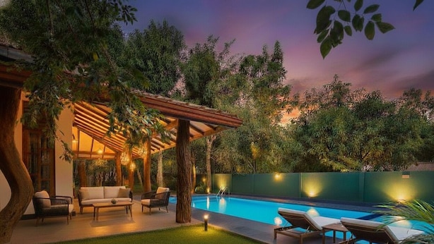 Un espace piscine doté de chaises longues et d’un coin salon abrité, entouré de verdure et surplombant le ciel nocturne.
