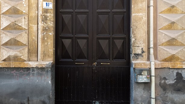 Eine schwarze, bogenförmige Tür mit einem dekorativen Metallwappen, eingebettet in eine beigefarbene Wand mit geometrischen Mustern.