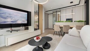 Ett modernt vardagsrum med en stor platt-TV, en soffa, en matplats med stolar och ett köksområde med kylskåp.