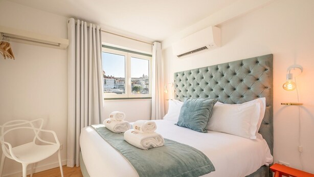Une chambre d’hôtel avec un grand lit, une fenêtre avec des rideaux, une chaise, une table de chevet et une lampe fixée au mur.