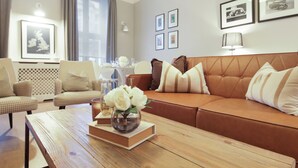 Ein modernes Wohnzimmer mit einem braunen Ledersofa, einem Holztisch und einem gemusterten Sessel. An den Wänden befinden sich Bilderrahmen und auf dem Tisch steht eine Vase mit Blumen.
