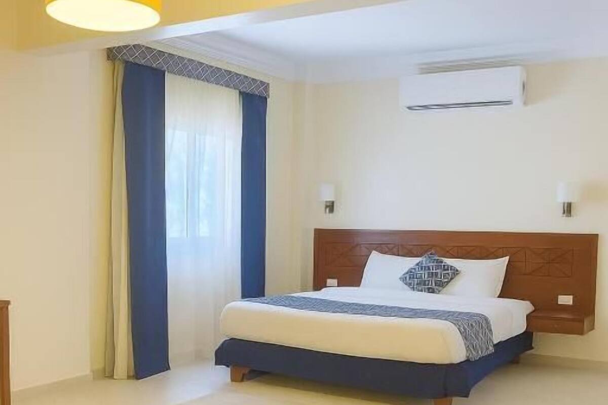 A hotel room with a bed, a bedside table, a lamp, and an air conditioning unit on the wall.