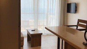 Een moderne hotelkamer met een houten eettafel, een kleine zithoek, een flatscreen televisie aan de muur en een groot raam met dunne gordijnen.