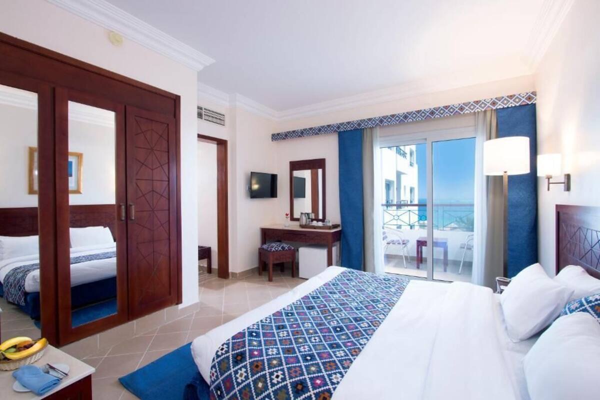 A hotel room with a large bed, a desk with a chair, a television, and a balcony with a view.