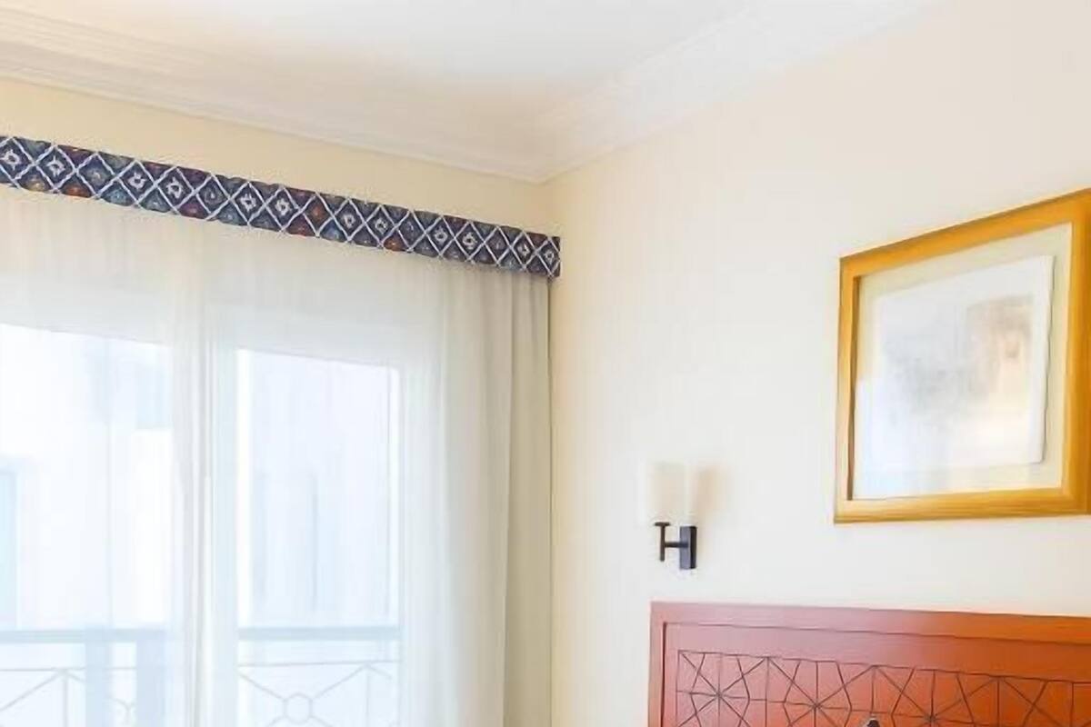 A hotel room with a bed, a nightstand, a wooden side table, a window with curtains, and a framed picture on the wall.