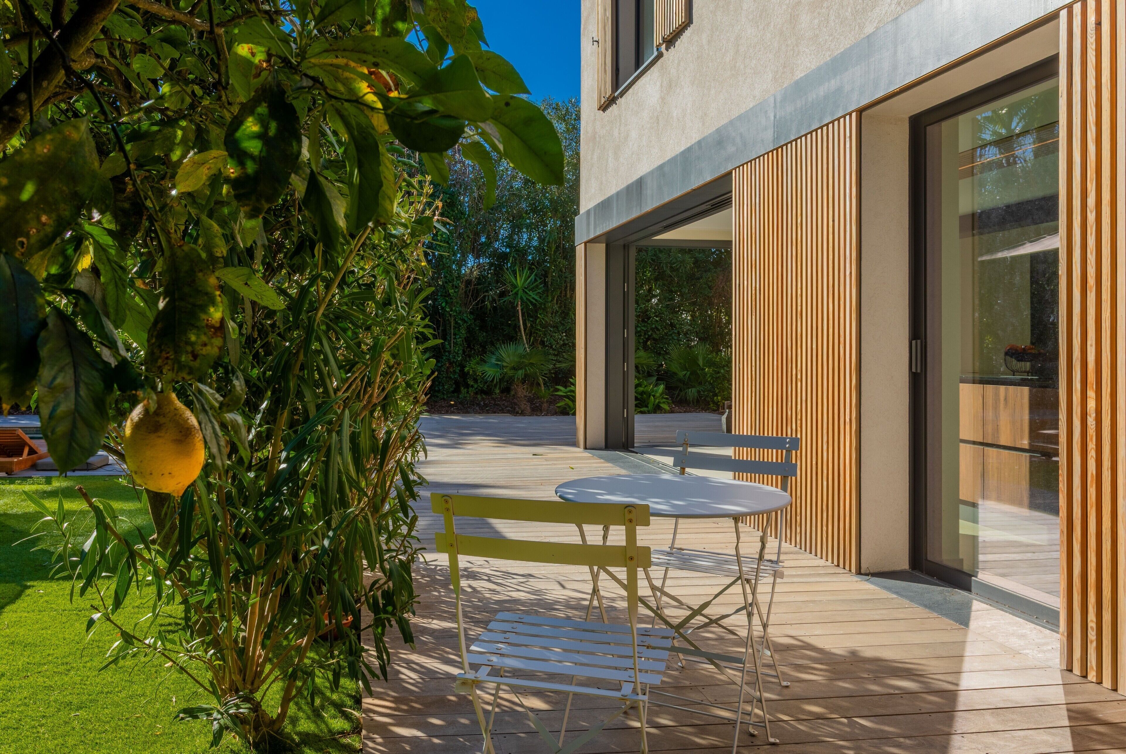 A modern outdoor patio with a wooden table and chairs, surrounded by greenery and a building with large windows.