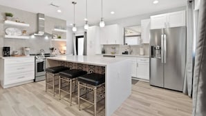Et moderne køkken med hvide skabe, en central ø med barstole, rustfrie stålapparater, hængende lysarmaturer og et backsplash med et plettet mønster.