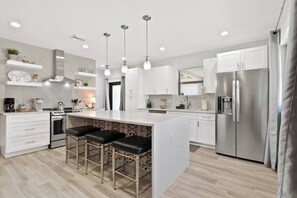 A modern kitchen with white cabinetry, a central island with bar stools, stainless steel appliances, pendant lighting, and a backsplash with a speckled pattern. - Newly Constructed Home near Downtown and Beaches (West Palm Beach)
