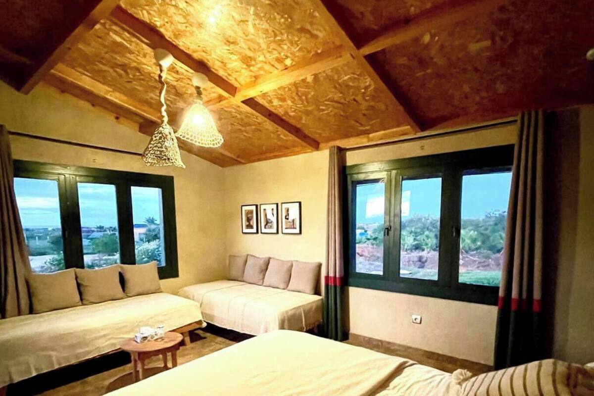 A room with two beds, a wooden ceiling, and large windows with curtains.