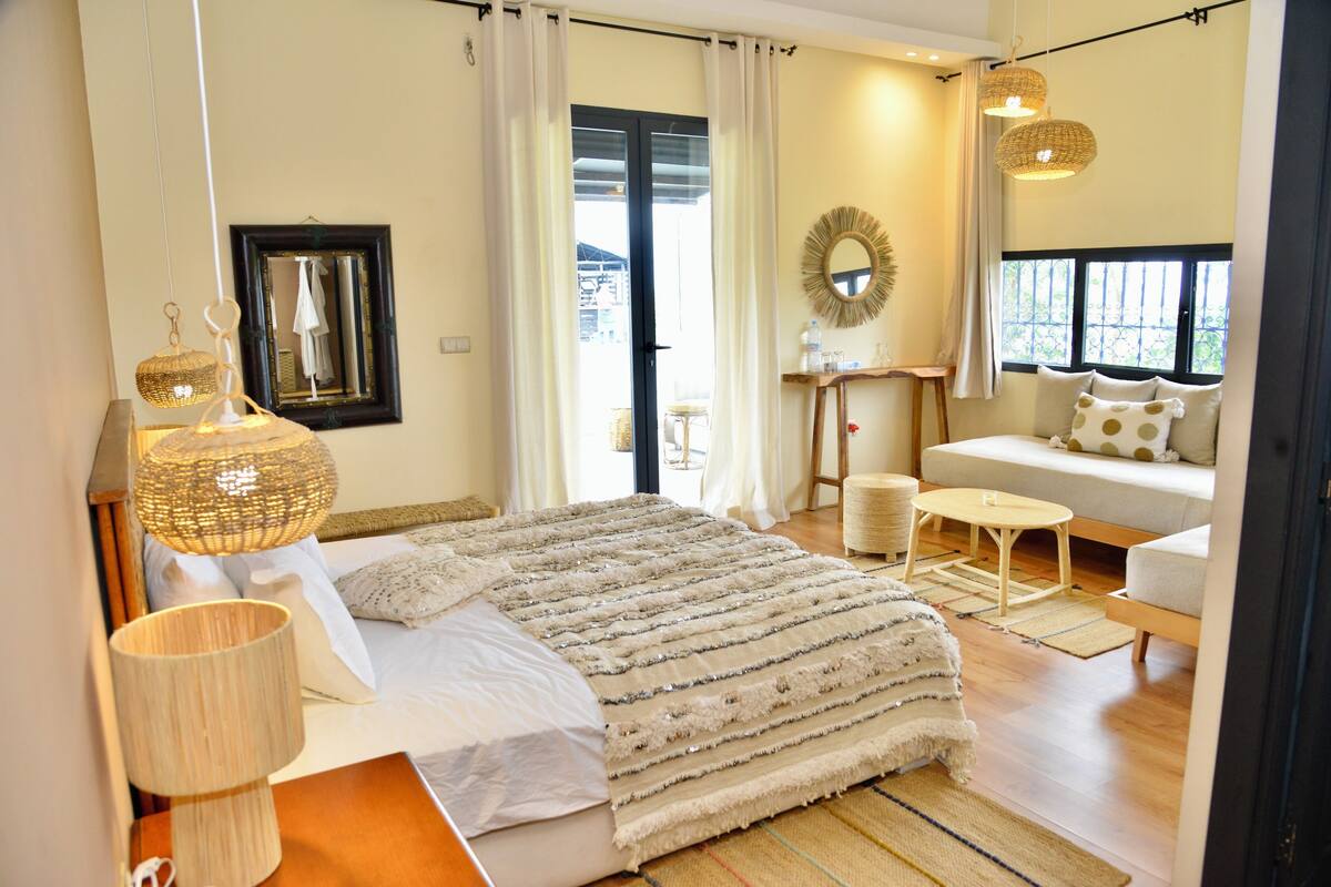 A bedroom with a bed, bedside table, lamp, and a view of the outside.