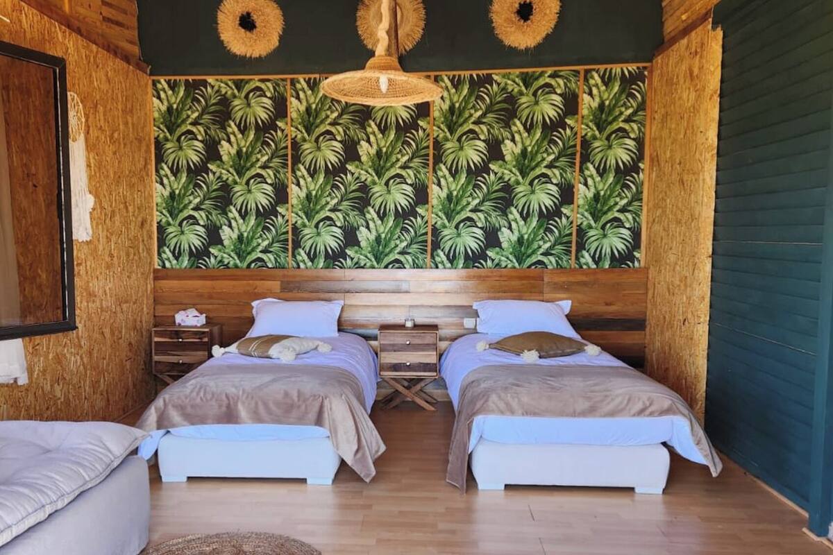 A room with two beds, a wooden ceiling, a tropical wallpaper, and a hanging light fixture.