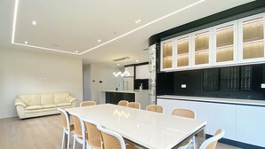 A modern kitchen with white cabinets, a large island, and a dining area with a white table and wicker chairs. - Koromeke Luxury 370sqm New Build– Ideal for Groups (Auckland)