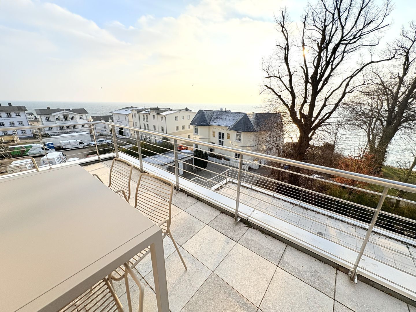 A balcony with a table and chairs, overlooking a waterfront with houses and a tree.