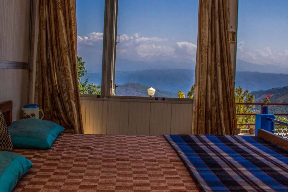 A bedroom with a checkered bedspread, a view of mountains through large windows, and a balcony with a railing.