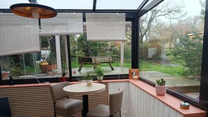 A modern indoor patio with a round table and two chairs, overlooking a garden with a wooden deck and potted plants. - Atipyk'Ododo (Saint-James)
