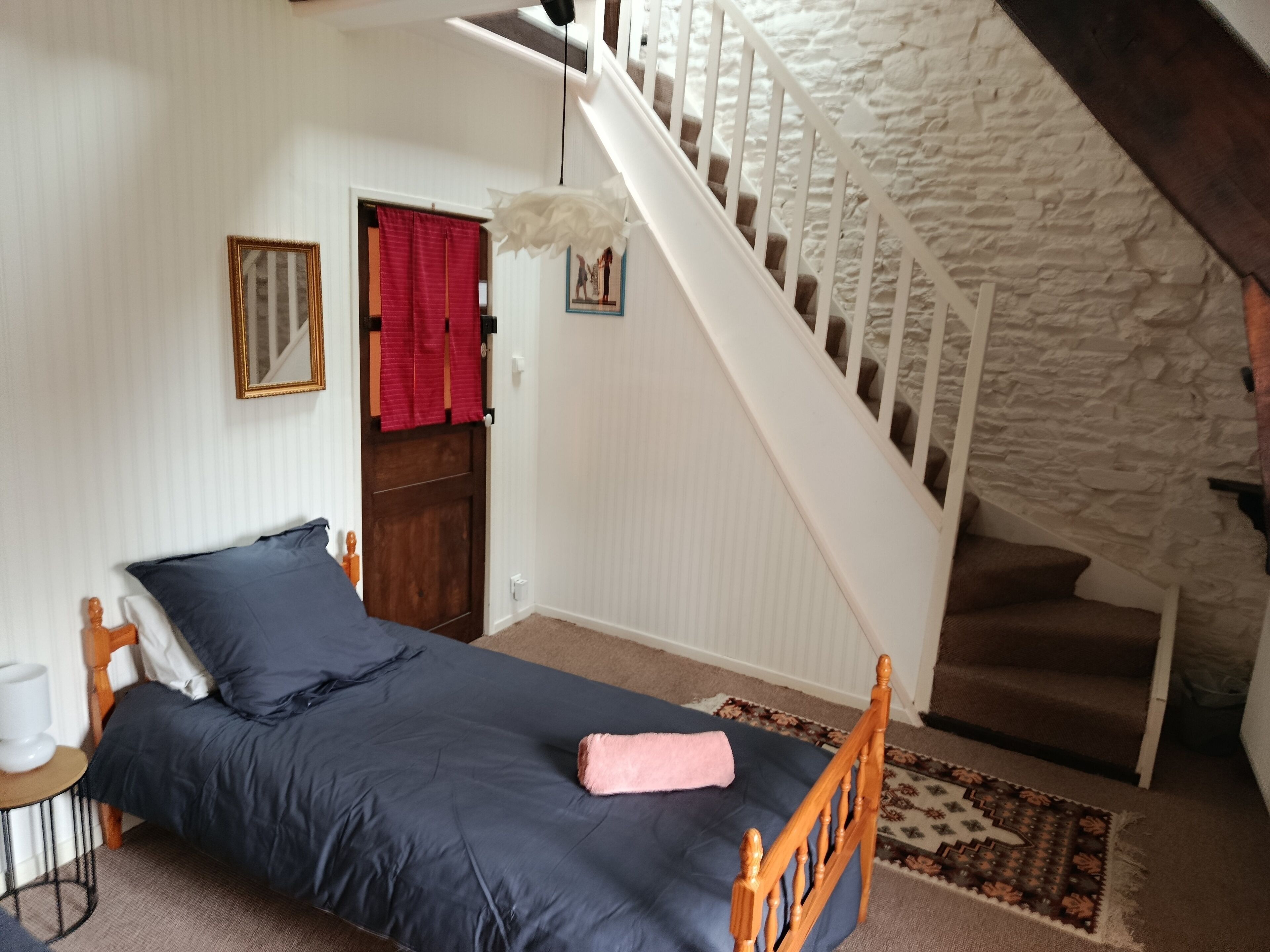Een slaapkamer met een enkel bed, een houten hoofdbord, een witte muur, een rood gordijn, een witte deur, een trap met een witte leuning en een kamerbreed tapijt met dessin.