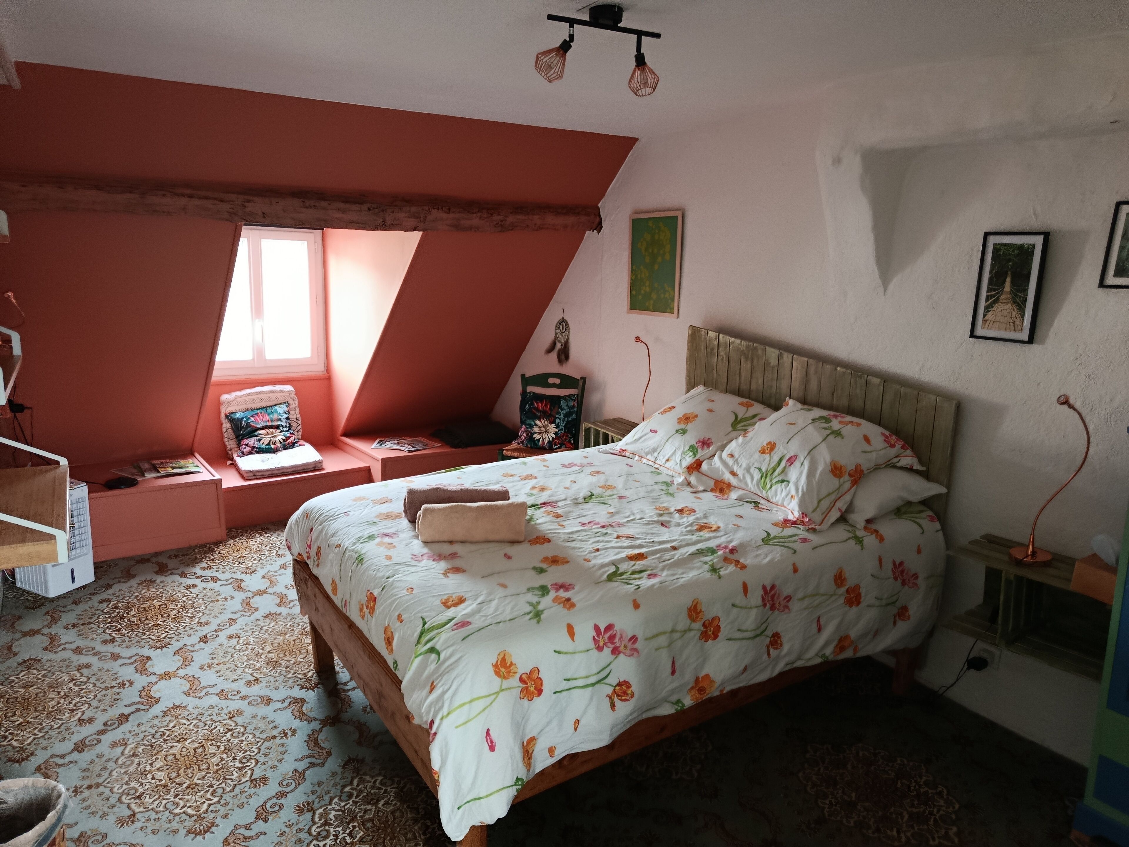 Un dormitorio con techo inclinado, una cama con ropa de cama floral, un sillón de lectura y una mesita de noche con lámpara.