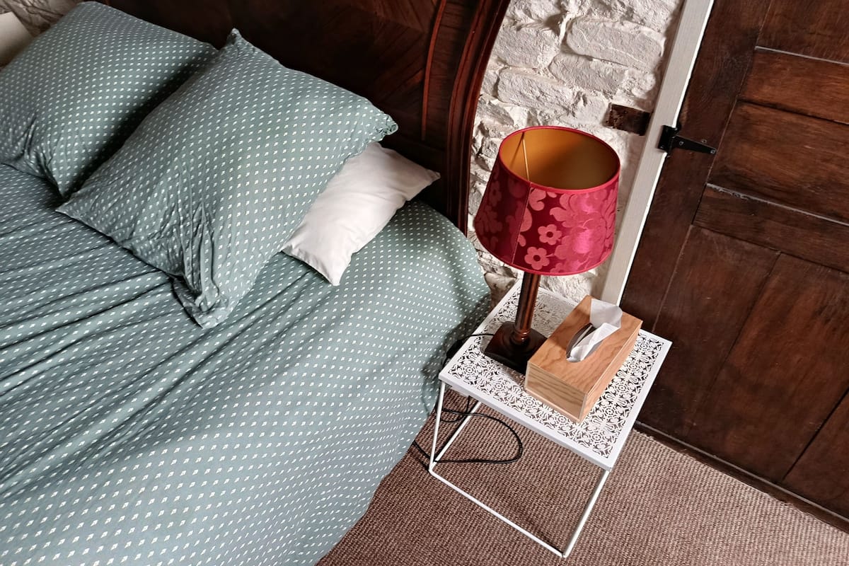 A neatly made bed with green bedding and a white pillow, a wooden bedside table with a lamp, and a white textured wall.
