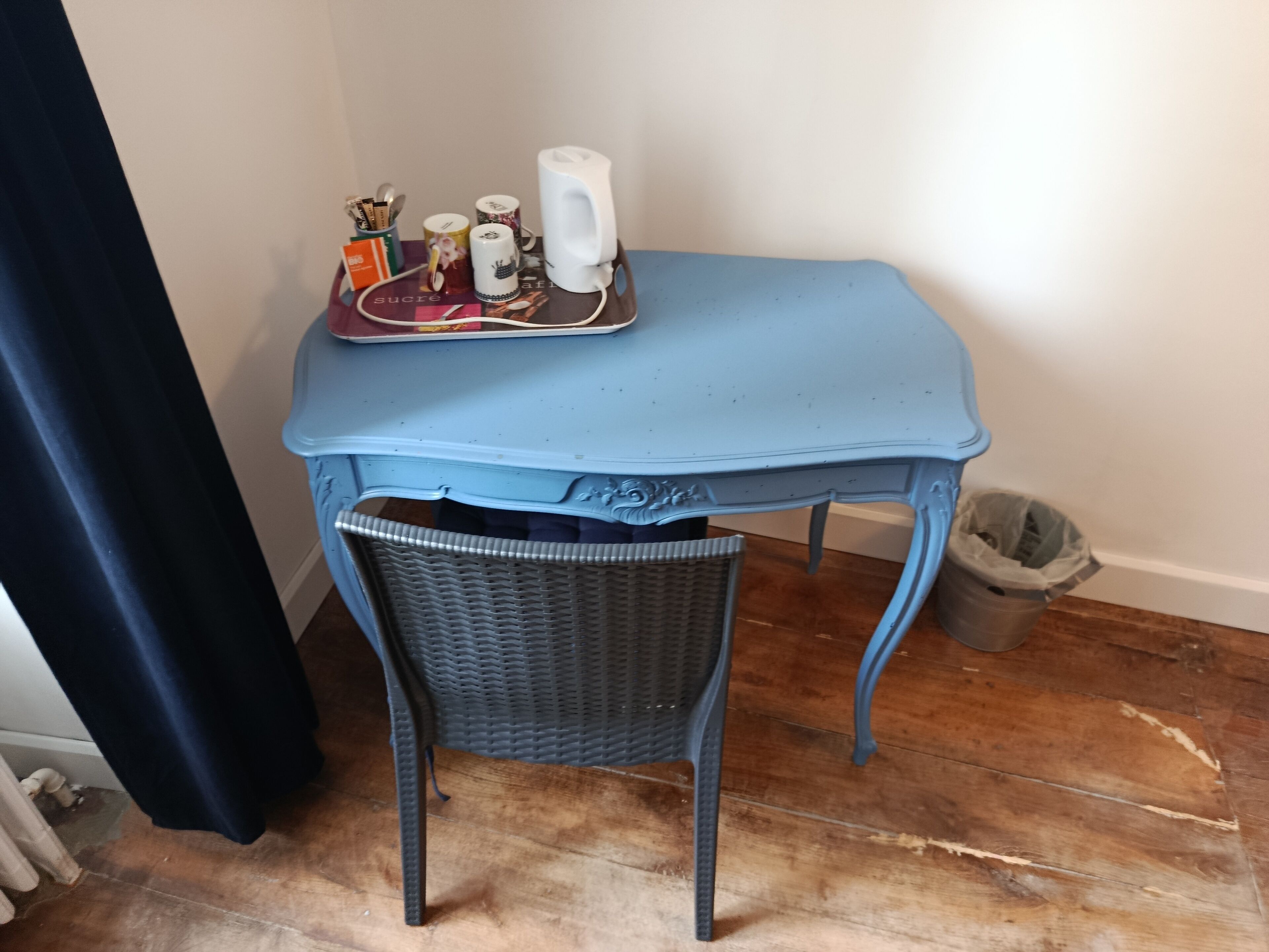 A blue table with a wicker chair, a tray with cups and a phone, and a trash can.