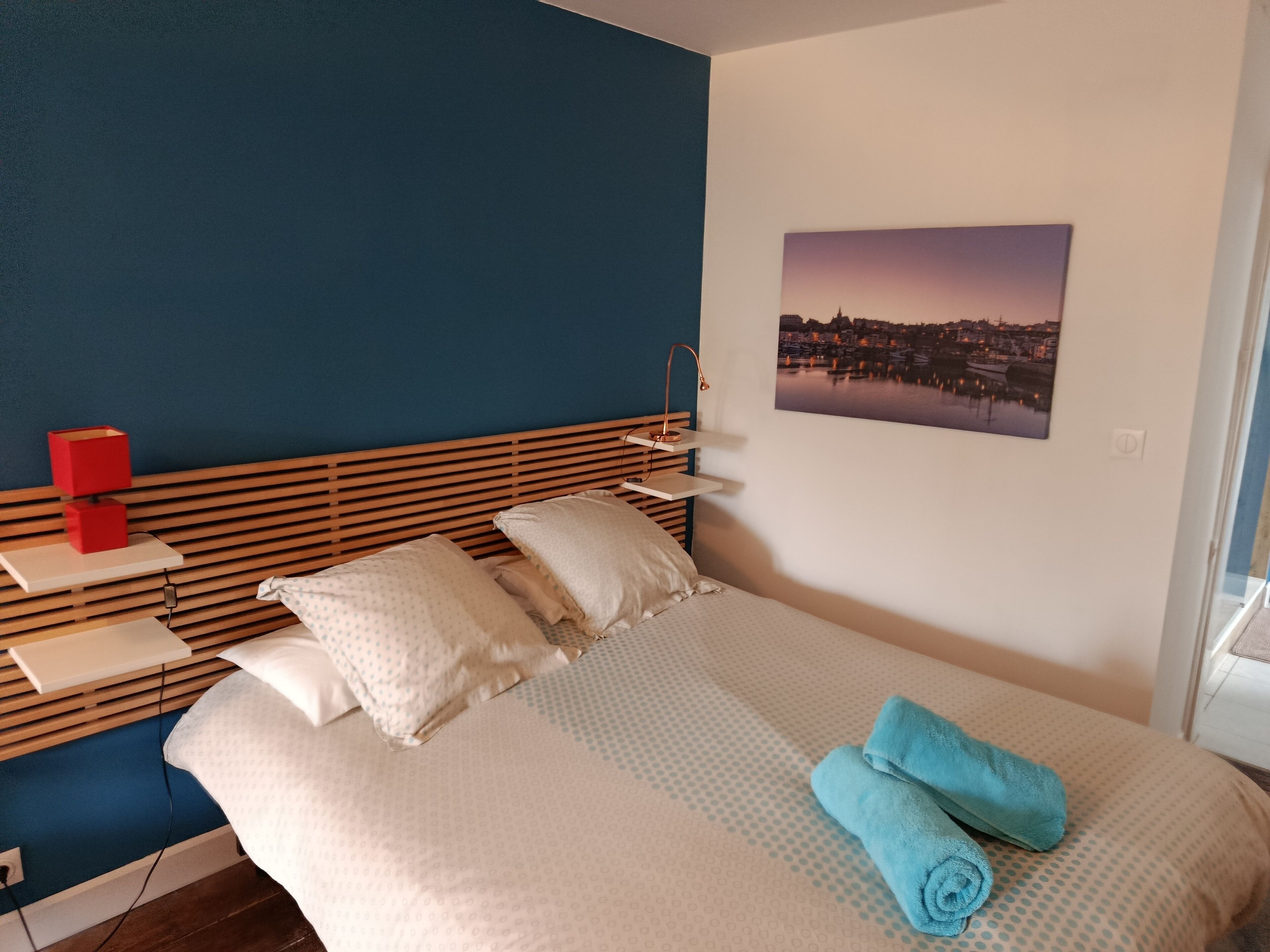 A neatly made bed with pillows and a folded towel, a bedside table with a red object, and a cityscape picture on the wall.