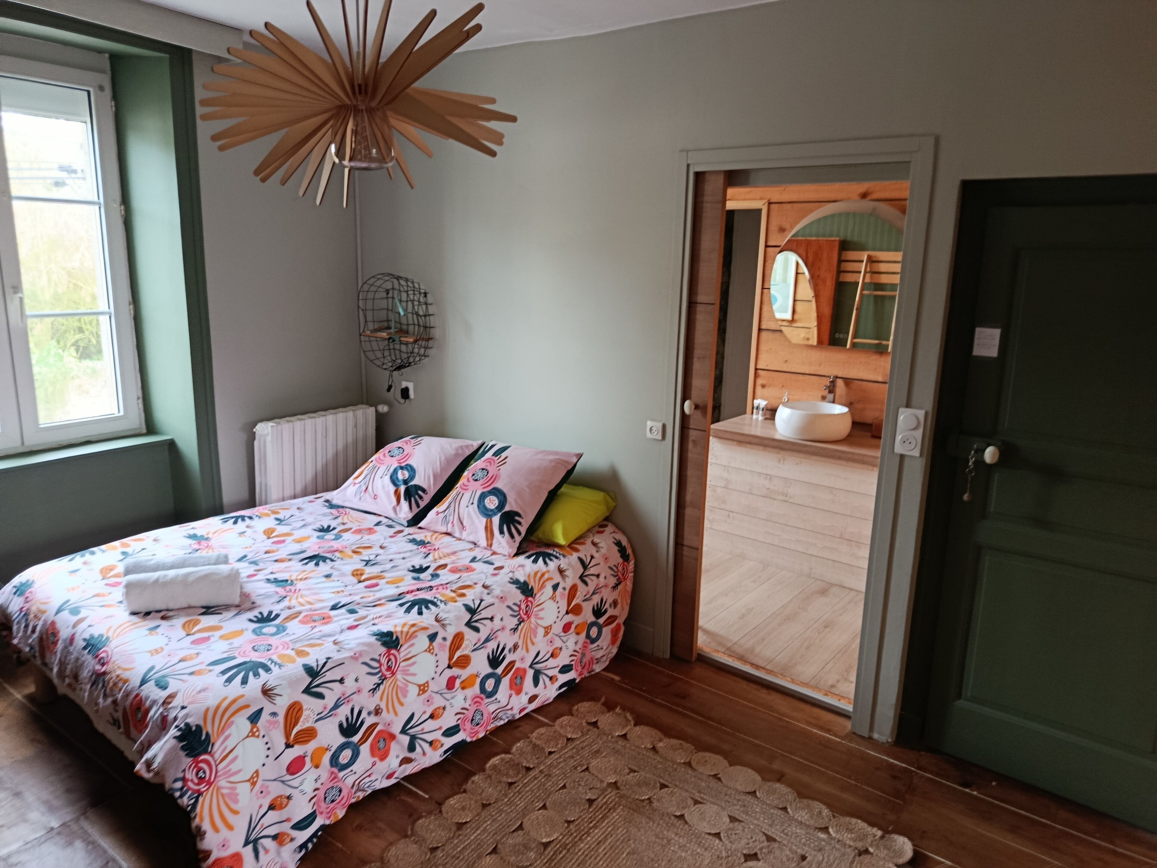 A bedroom with a floral bedspread, a wooden floor, a green door, and a view into the bathroom.