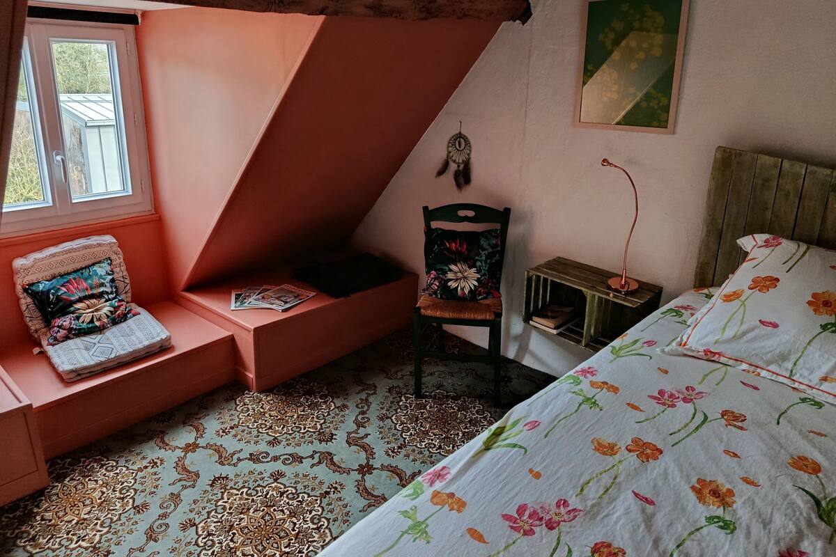 A cozy room with a sloped ceiling, a bed with floral bedding, a chair with a floral cushion, a small table with a lamp, and a dreamcatcher on the wall.