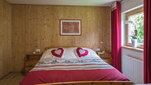 A bedroom with a wooden paneled wall, a bed with heart-shaped pillows, a nightstand with a lamp, and a window with red curtains. - CUCOL HUT (Trentino-Alto Adige)