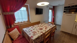 A cozy room with a wooden bench, a table with a patterned tablecloth, a TV mounted on the wall, and a kitchenette with a stove and oven. - CUCOL HUT (Trentino-Alto Adige)