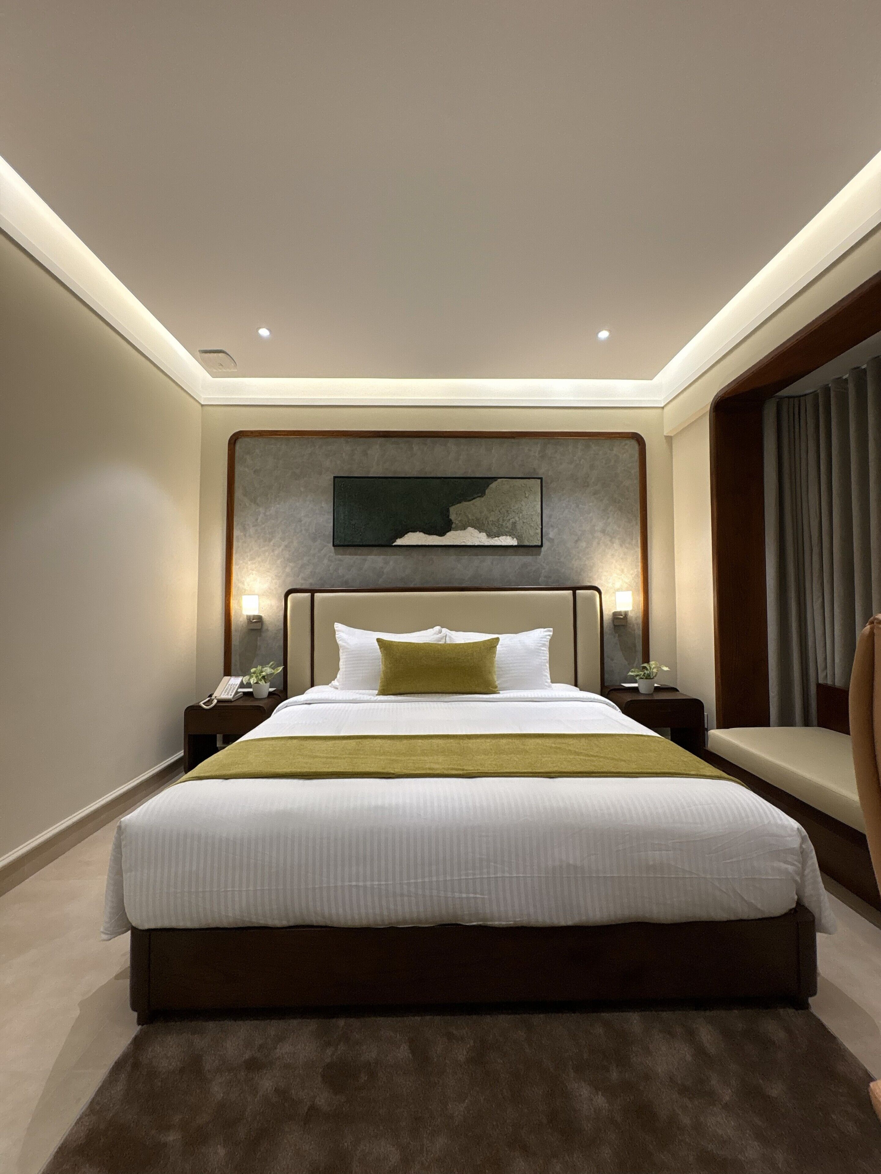 A modern hotel room with a large bed, bedside tables, a nightstand, a chair, and a painting on the wall.