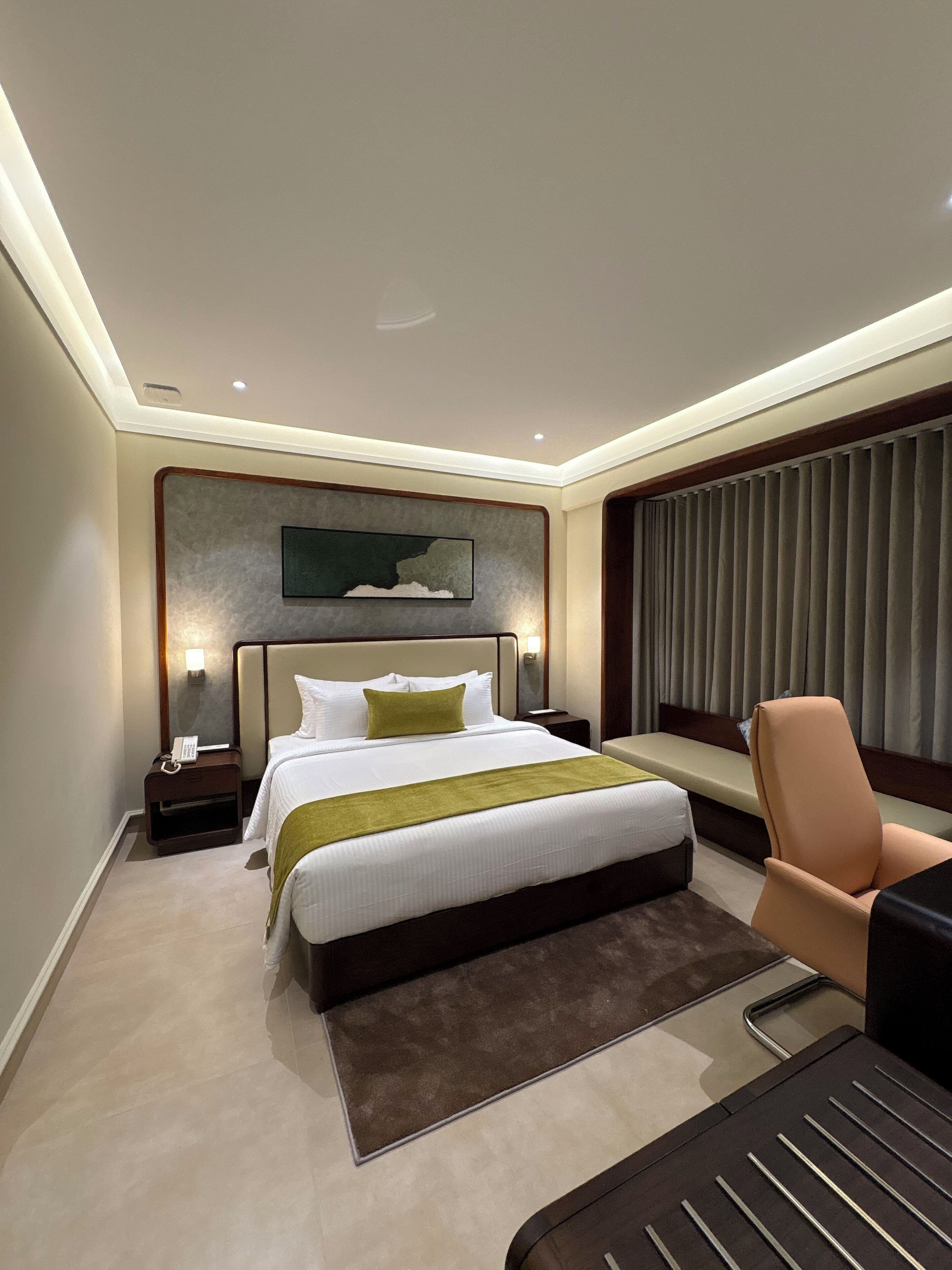 A modern hotel room with a large bed, a bedside table with a lamp, a chair, and a view of the city through the window.