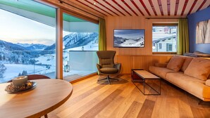 Un soggiorno moderno con pavimento in legno, un divano, una poltrona e un tavolo con una candela. Grandi finestre offrono vista su montagne innevate.