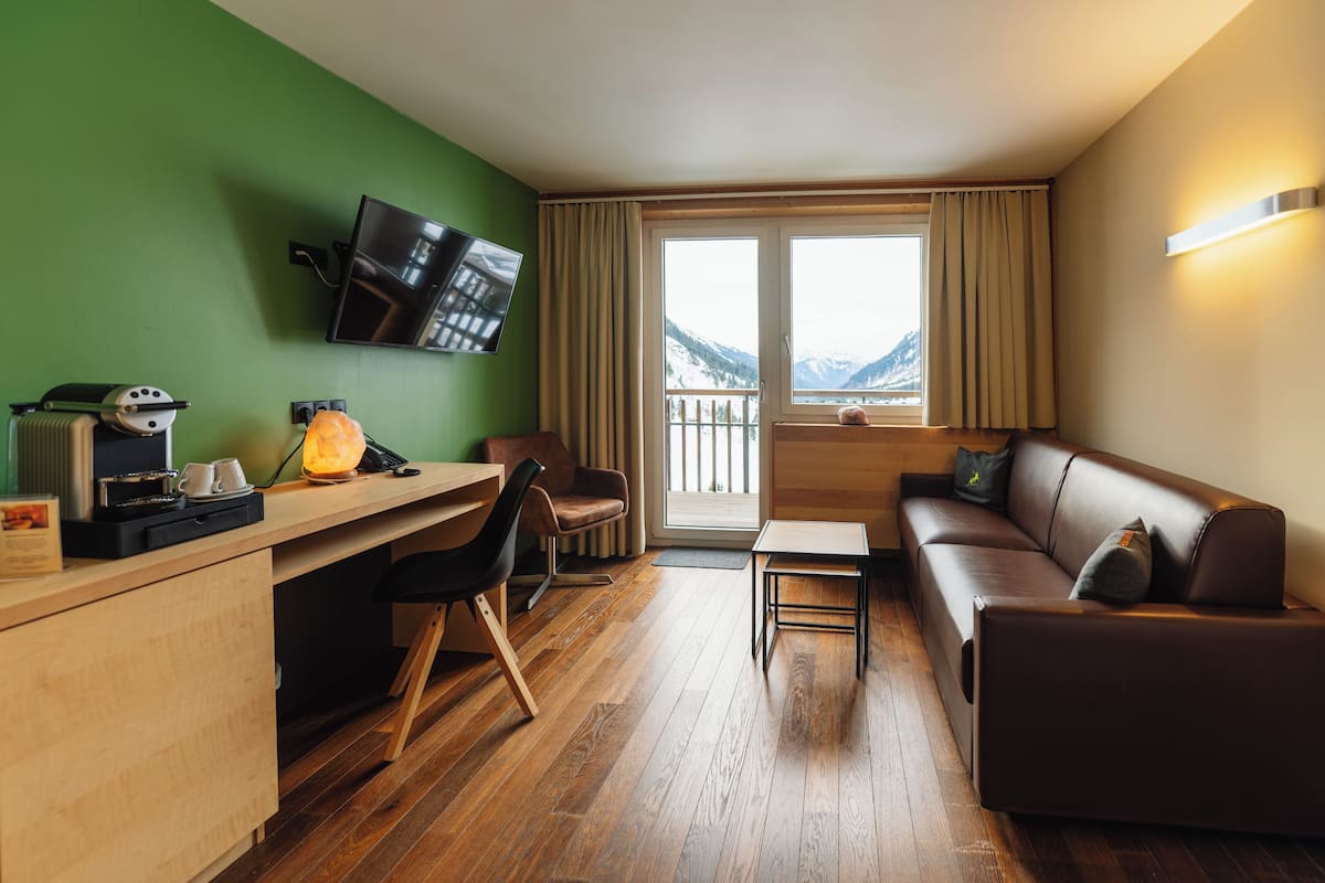 Ein modernes Hotelzimmer mit einem Holztisch, einer braunen Ledersofa und einem an der Wand befestigten Fernseher. Durch das Fenster ist eine Bergblick zu sehen.