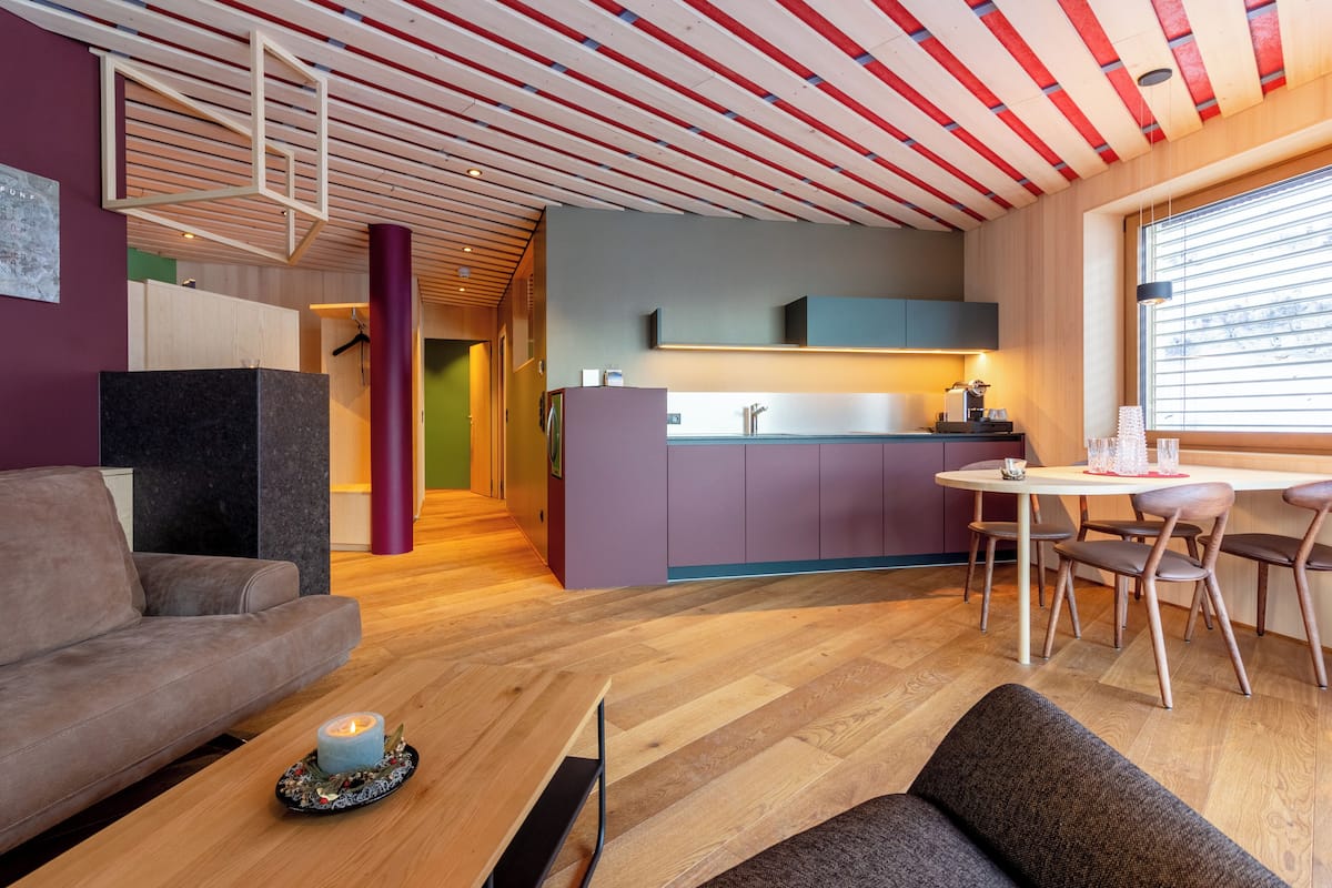 Ein modernes Wohnzimmer mit Holzboden, einer Couch, einem Esstisch mit Stühlen und einer Küchenzeile mit lilafarbenen Schränken.