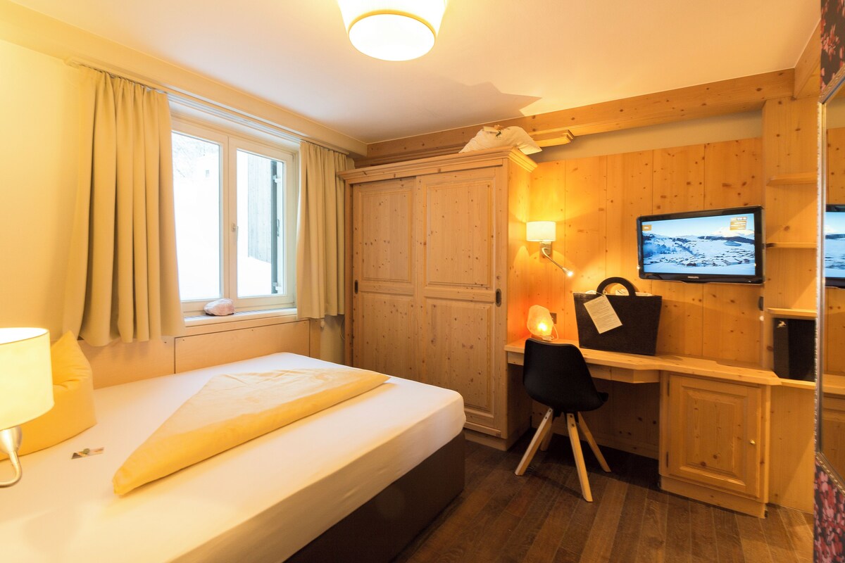 Habitación de hotel con cama, escritorio, silla, televisor y paredes de paneles de madera.