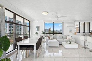 A modern living room with a dining area, large windows, and a view of high-rise buildings. - Cosy Ocean Views Apartment w/cinema at night. Pet-Friendly! (Surfers Paradise)