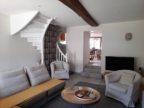 Ein modernes Wohnzimmer mit einer Couch, Sesseln, einem Couchtisch und einem Fernseher. Eine Treppe führt zu einer oberen Ebene mit einem Essbereich.