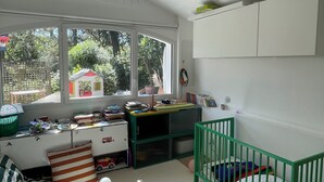 Ein Kinderzimmer mit einem Gitterbett, einem Schreibtisch und einem Fenster mit Blick auf Grünflächen.