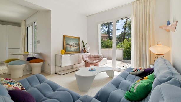 Ein modernes Wohnzimmer mit einer Couch, einem Sessel und einem Couchtisch. An der Wand hängt ein Gemälde und ein Fenster mit Vorhängen.